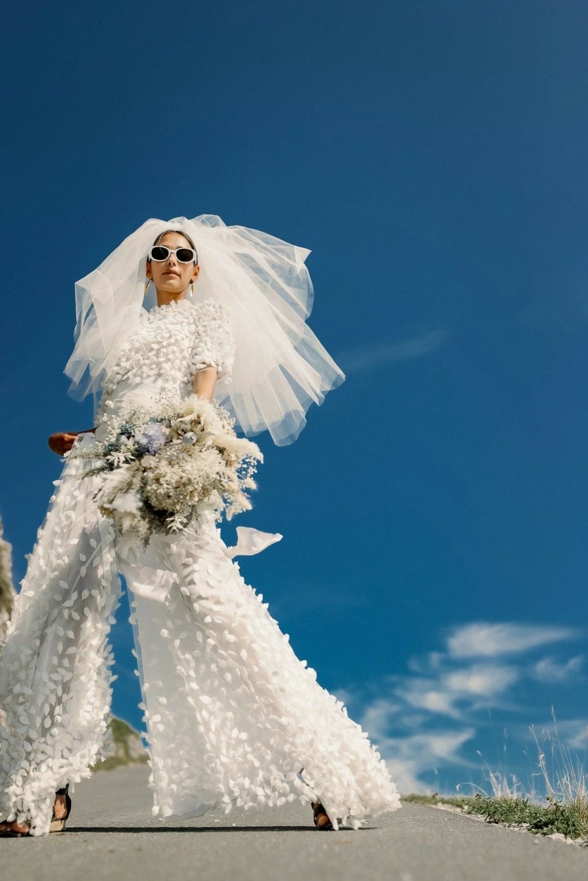 Noiva sem vestido, em macacão branco com véu e buquê em céu azul.