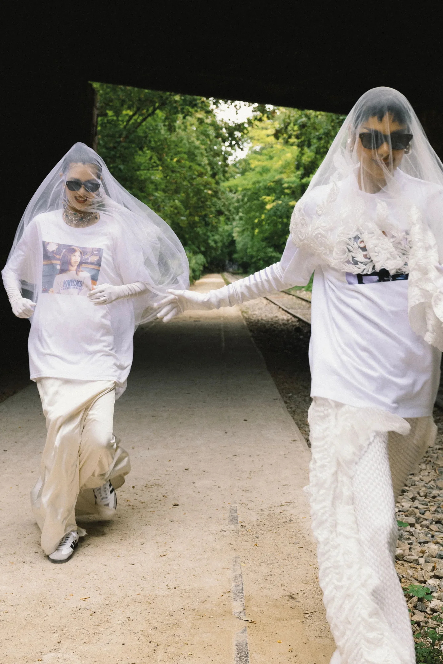 Duas pessoas em túnicas brancas e véus transparentes correm em um túnel arborizado, usando óculos escuros. Noiva sem vestido