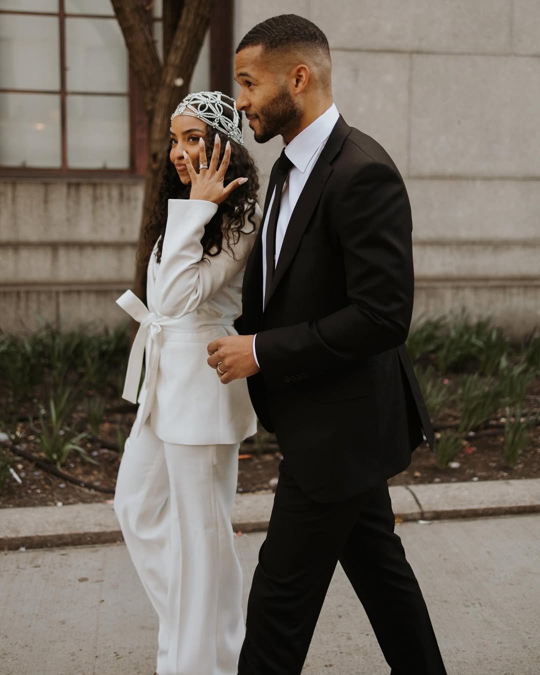 Casal caminha. A noiva, em branco, usa um terno elegante e mostra a aliança, sorrindo. Noiva sem vestido