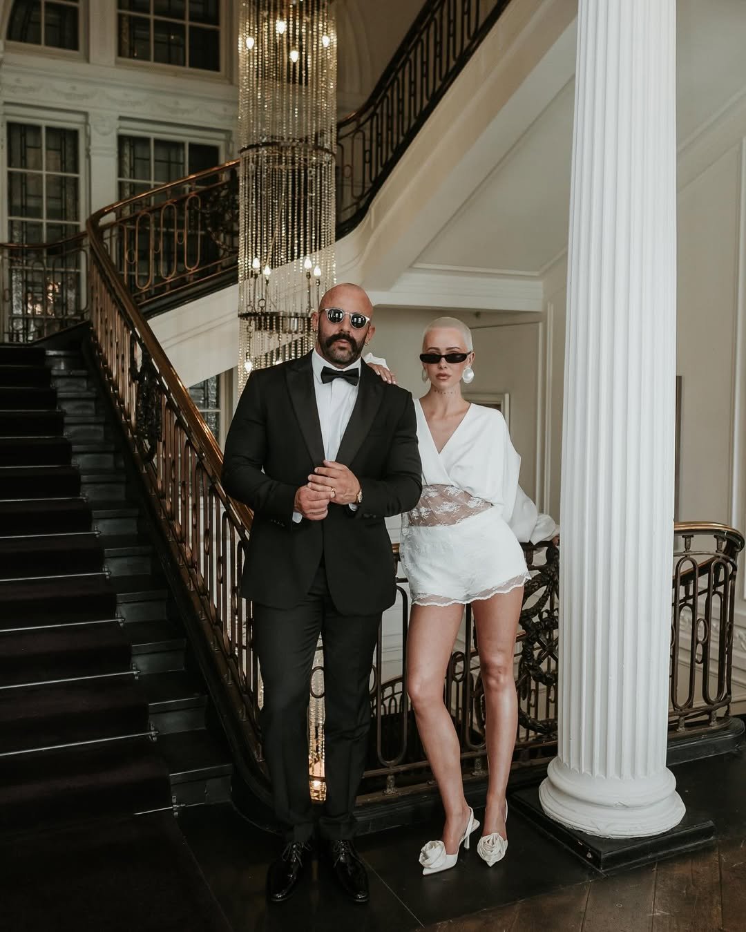 Casal elegante posa em escadaria luxuosa; mulher usa traje branco alternativo nupcial sem vestido. Noiva sem vestido