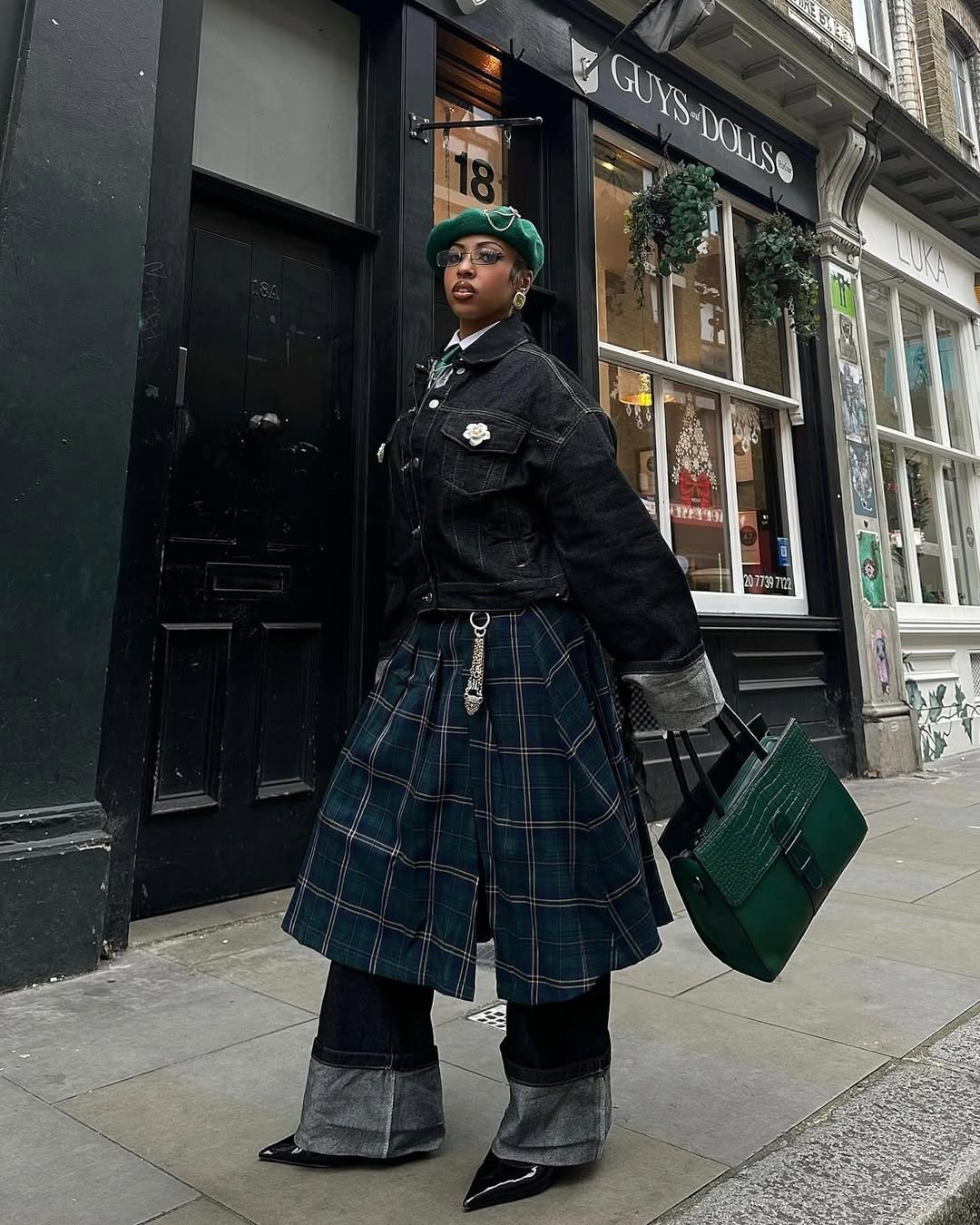 Pessoa usando casaco preto e boina, combinando saia xadrez estilo vestido sobre calça escura larga.