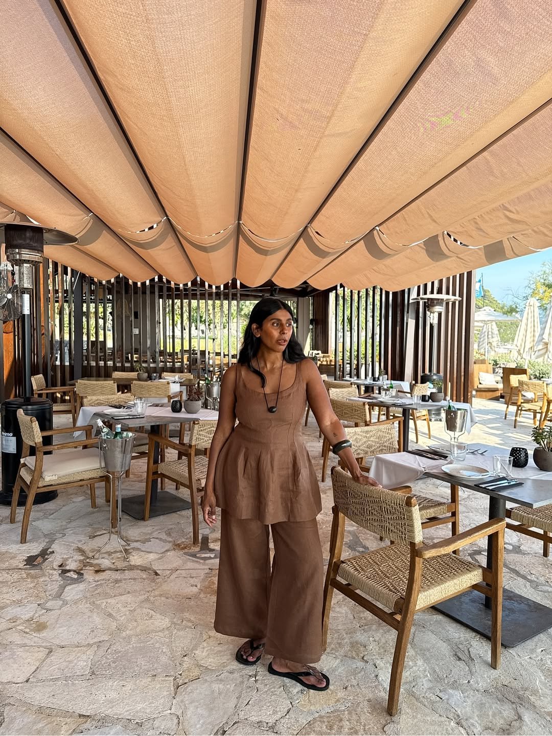 Mulher em um restaurante ao ar livre usa vestido e calça marrom sob teto de lona beige.