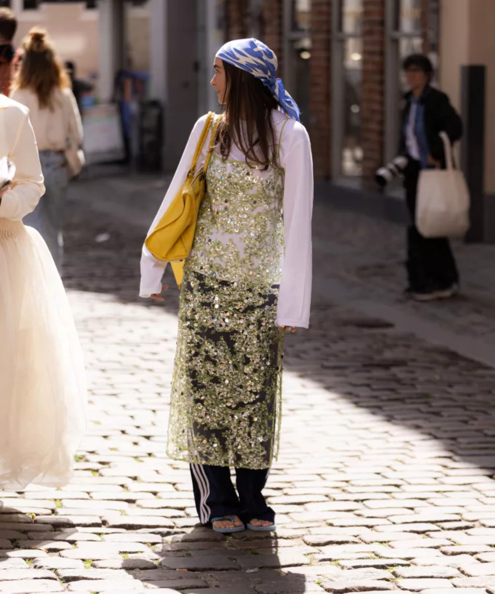 Mulher com lenço azul, vestido com lantejoulas sobre calça preta listrada, segura bolsa amarela em rua de paralelepípedos.