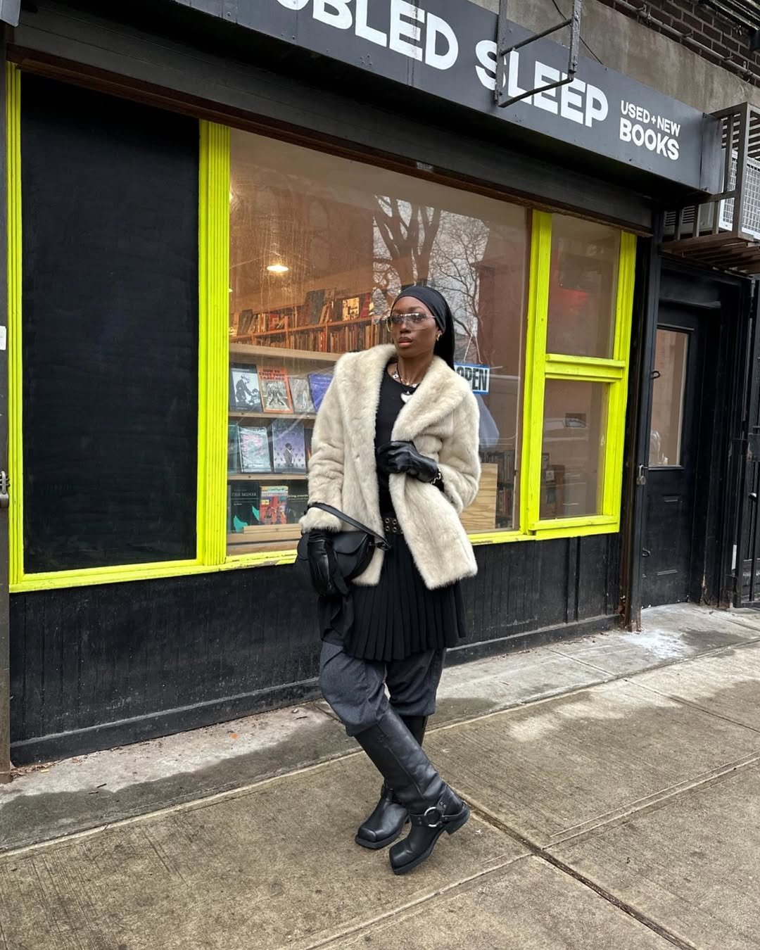 Pessoa com vestido preto por cima da calça, usando um casaco de pelos e uma bota preta.