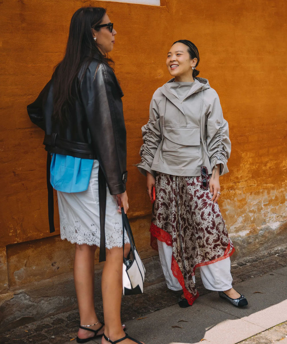 Duas mulheres conversam; uma com jaqueta de couro e vestido, outra com calça e blusa cinza.
