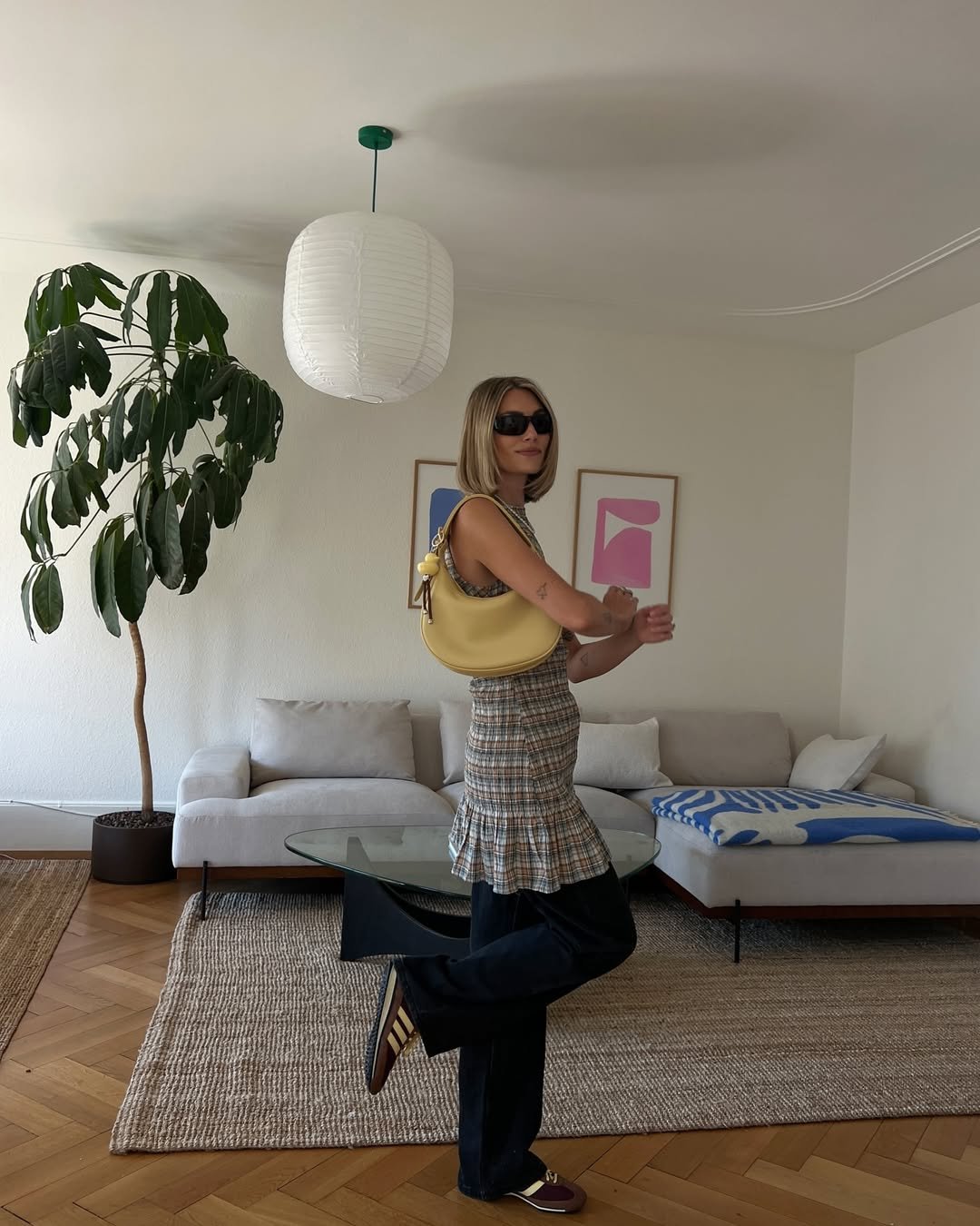 Pessoa usa vestido e calça preta, bolsa amarela. O ambiente é uma sala de estar com sofá e planta.