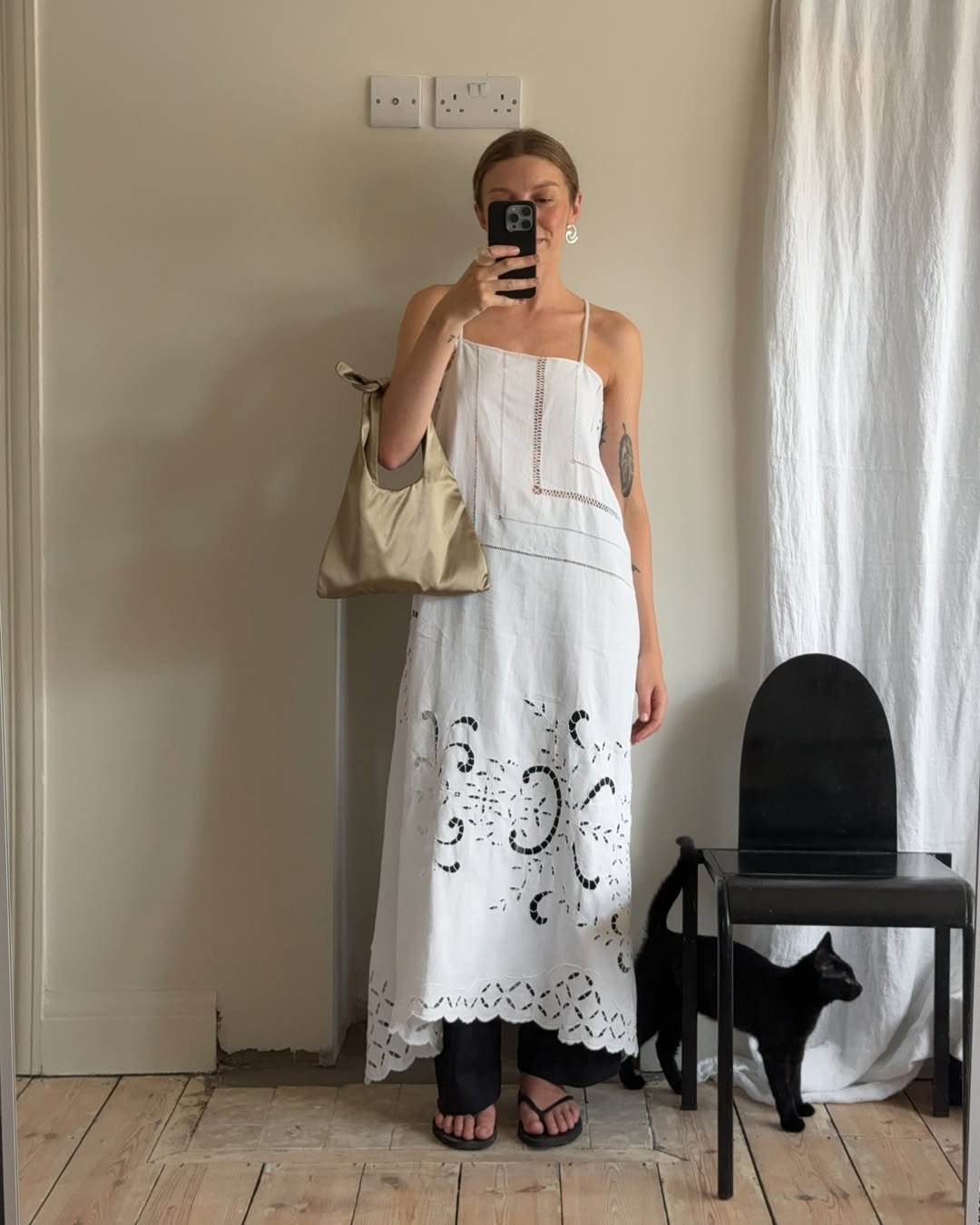 Pessoa usando vestido branco com detalhes vazados sobre calça preta e segurando bolsa bege. Gato preto ao lado.