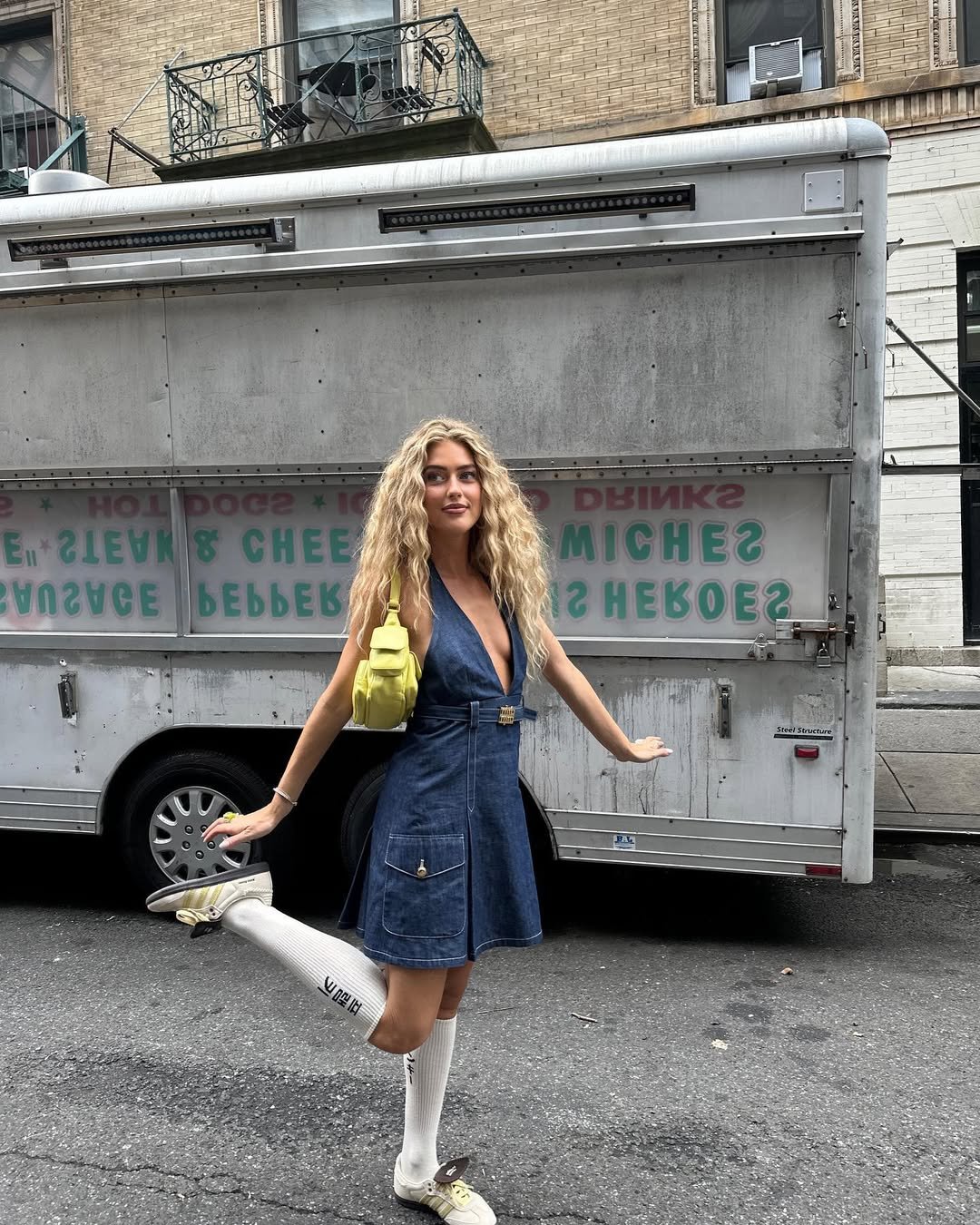 Pessoa posando na rua com vestido jeans, mochila amarela e tênis, em frente a um caminhão de comida.