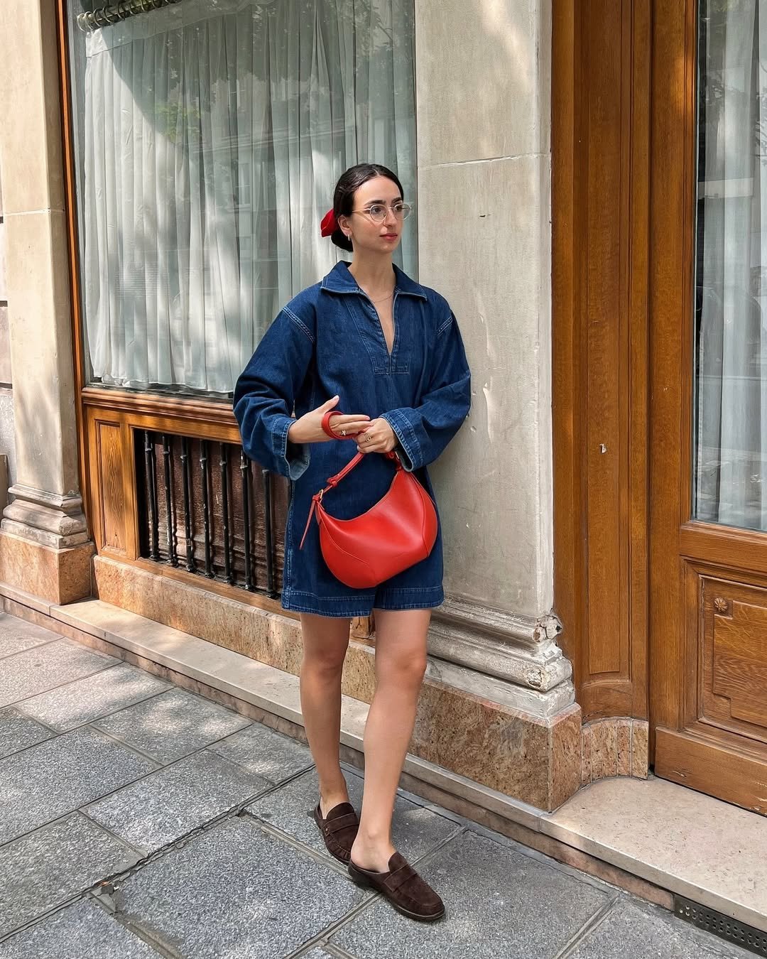 Pessoa usando vestido jeans largo, bolsa vermelha e sapatos marrons, parada em frente a uma janela com cortina branca.