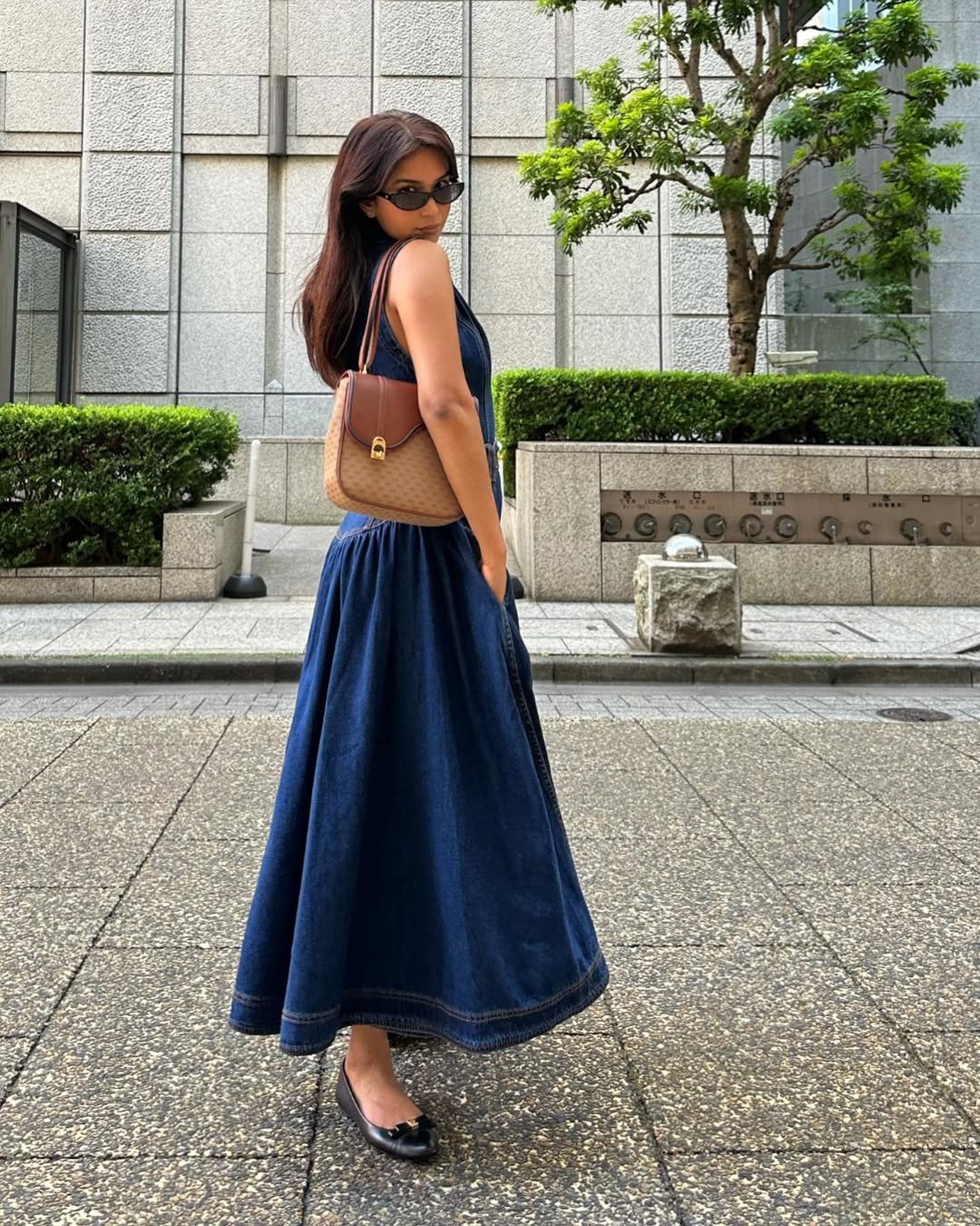 Pessoa de óculos escuros usa vestido jeans longo e bolsa marrom em rua arborizada.
