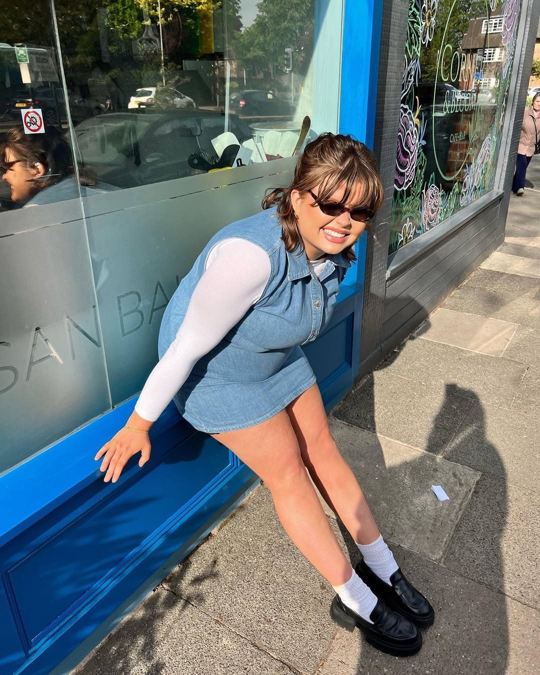 Pessoa sorrindo, inclinada em frente a uma janela. Ela veste um vestido jeans sobre blusa branca.