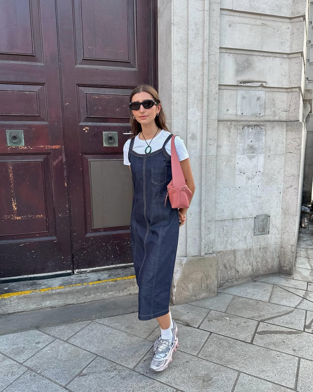 Mulher usando vestido jeans sobre camiseta branca, óculos escuros e bolsa rosa, em frente a uma porta de madeira.