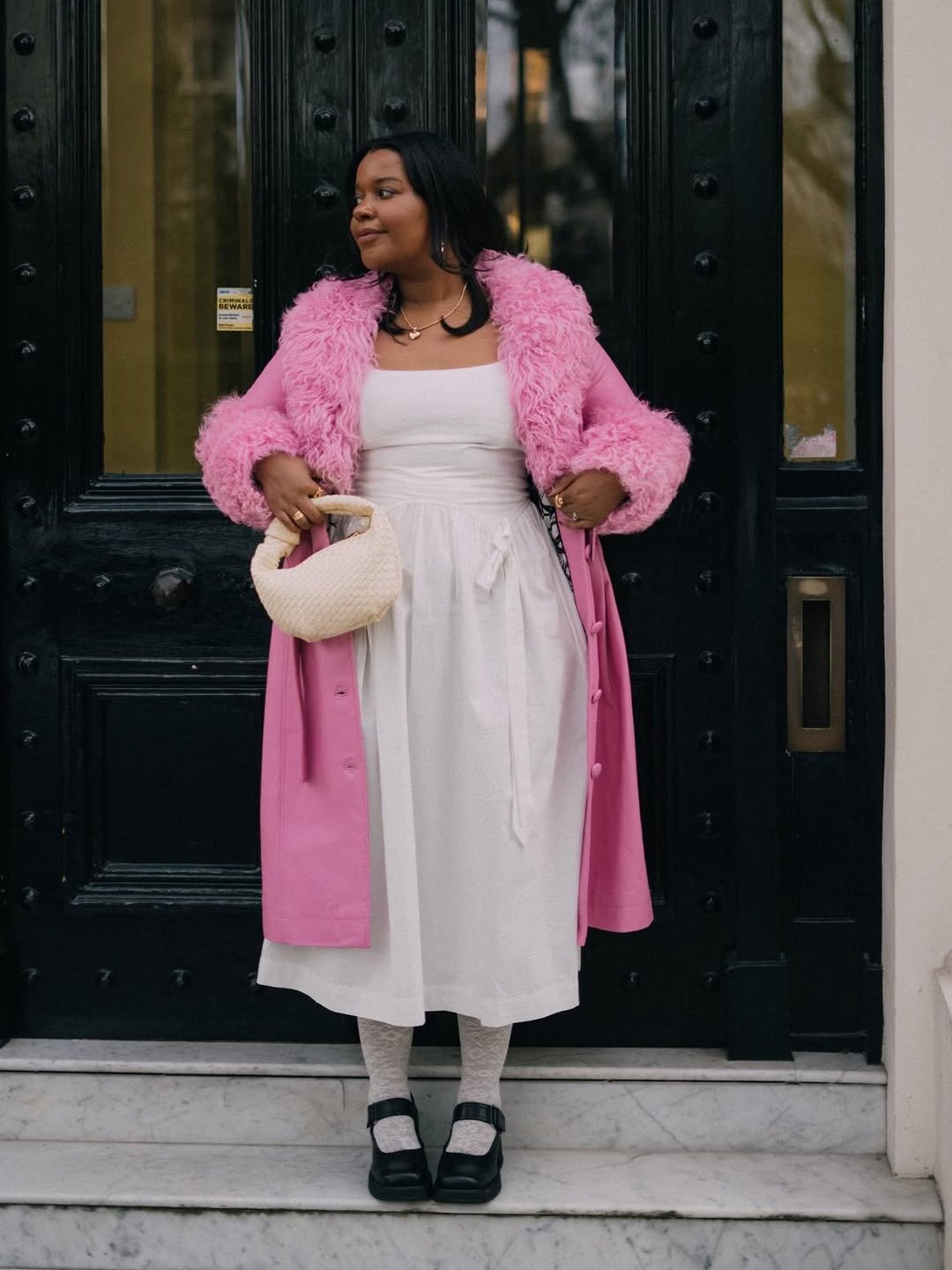 Mulher com casaco rosa e vestido branco médio, segurando bolsa, em frente a porta preta.