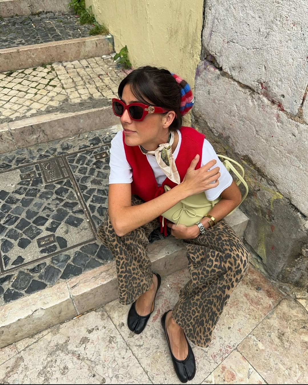 Mulher sentada em escada, usando óculos vermelhos e segura bolsa clara. Veste calça com estampa animal e colete vermelho.