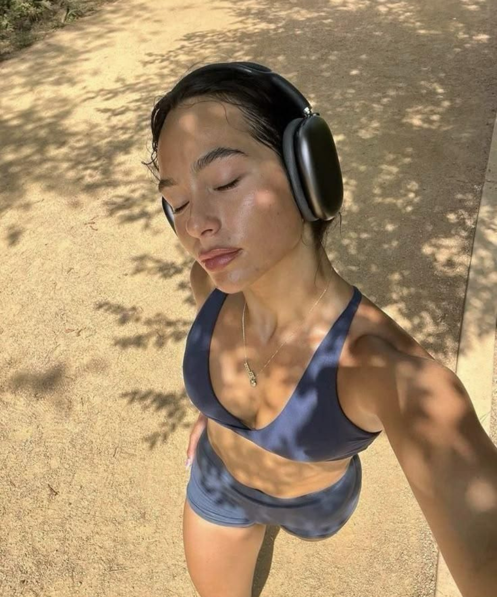 Jovem de olhos fechados, com fones de ouvido, em roupa de treino azul sob sombras de árvores.