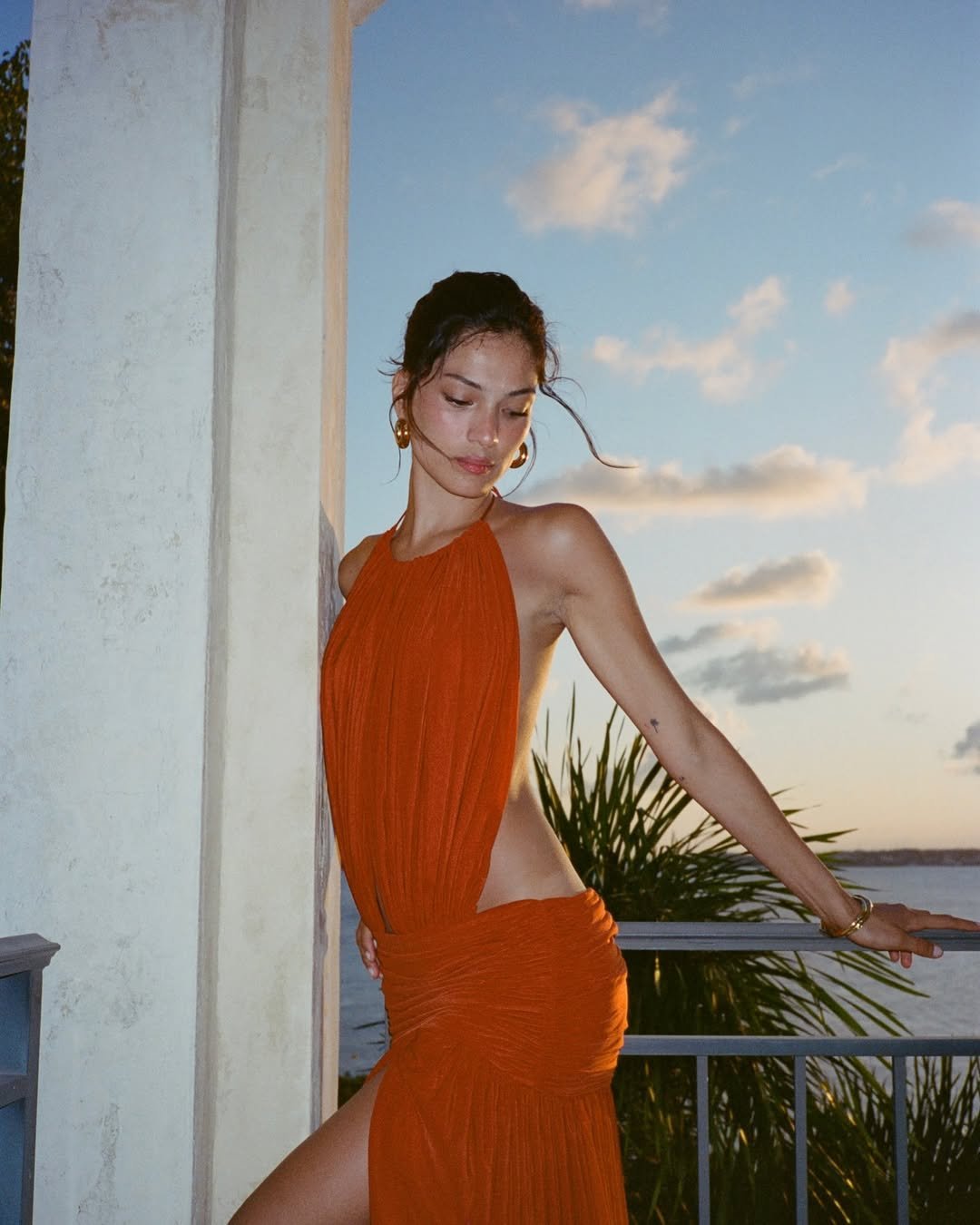 Mulher em vestido laranja posa em sacada ao pôr do sol, evocando elegância e estilo maximalista italiano.