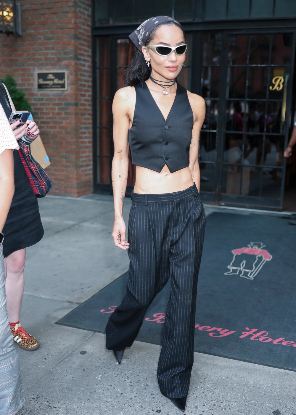 Zoë Kravitz com colete preto, calça de listras e lenço na cabeça, usando óculos de sol.