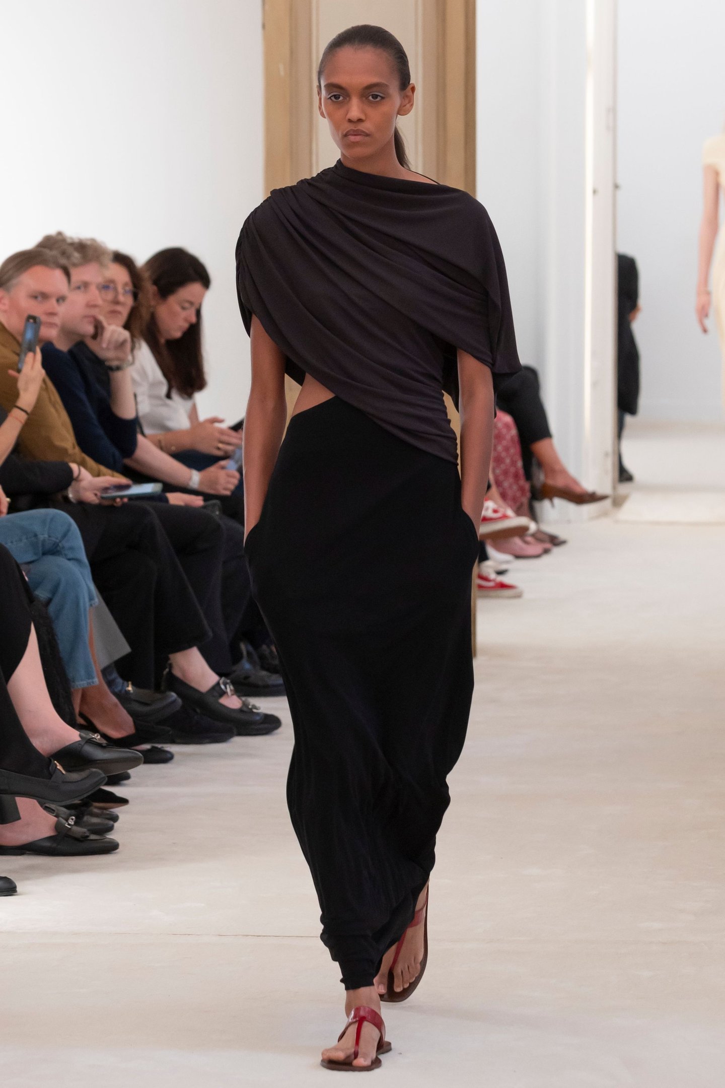 Desfile Milan Fashion Week SS26: modelo em vestido preto drapeado com sapatos vermelhos. Público atento ao fundo.
