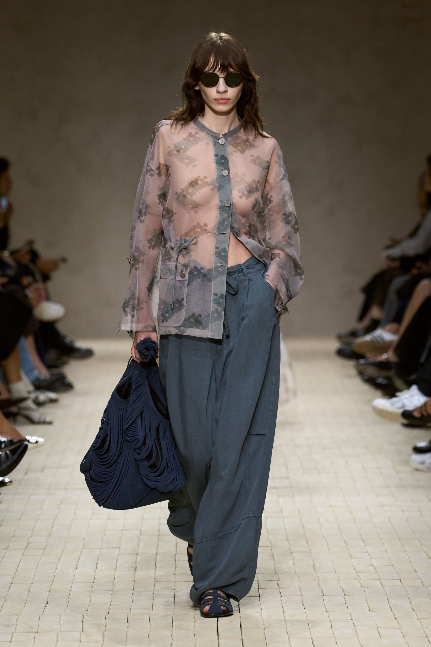 Modelo em passarela vestindo blusa transparente e calça azul, carregando bolsa azul durante a Milan Fashion Week SS26.
