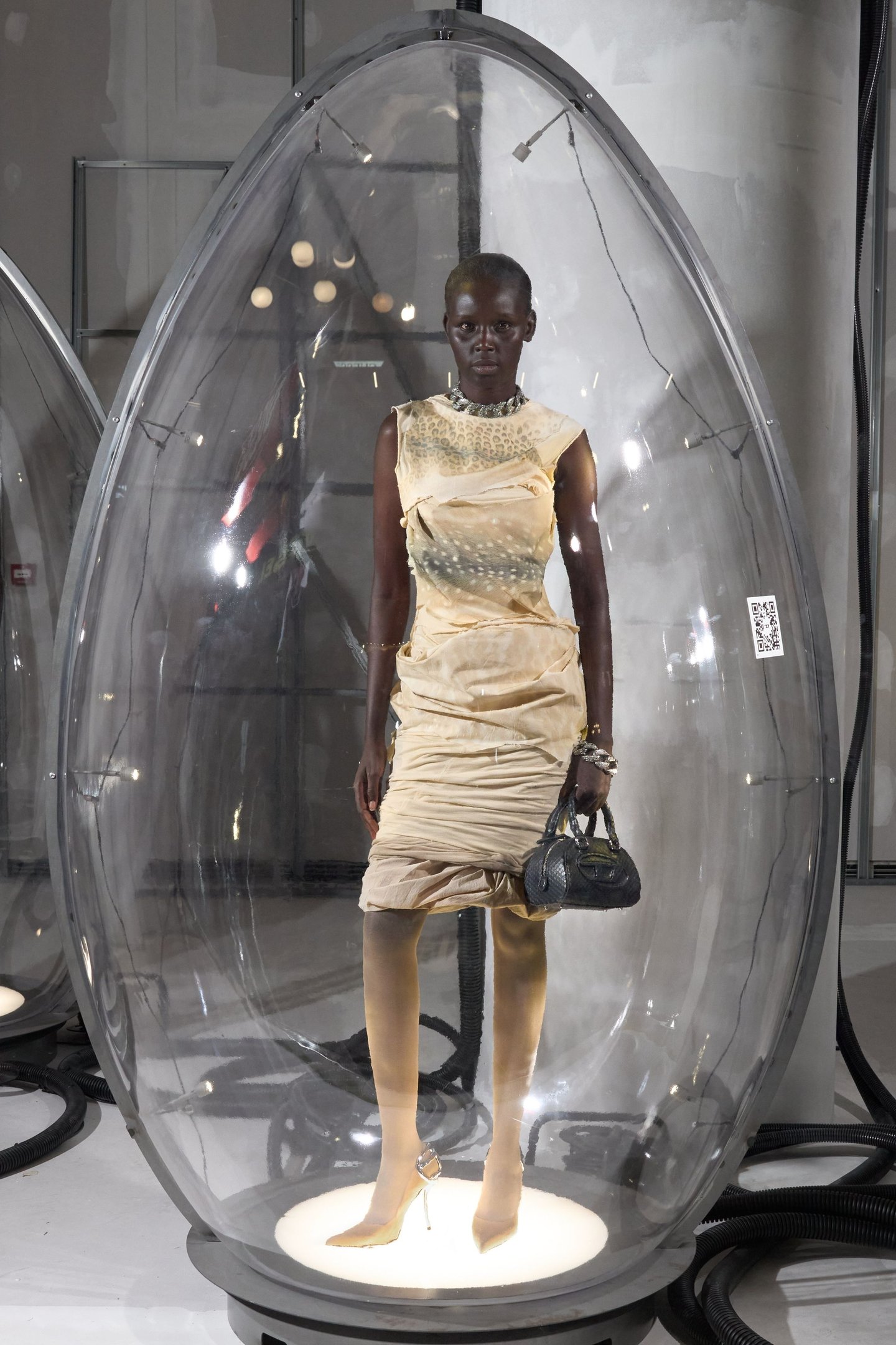 Modelo em vestido bege dentro de uma cápsula transparente na Milan Fashion Week SS26. Ela segura uma bolsa preta.