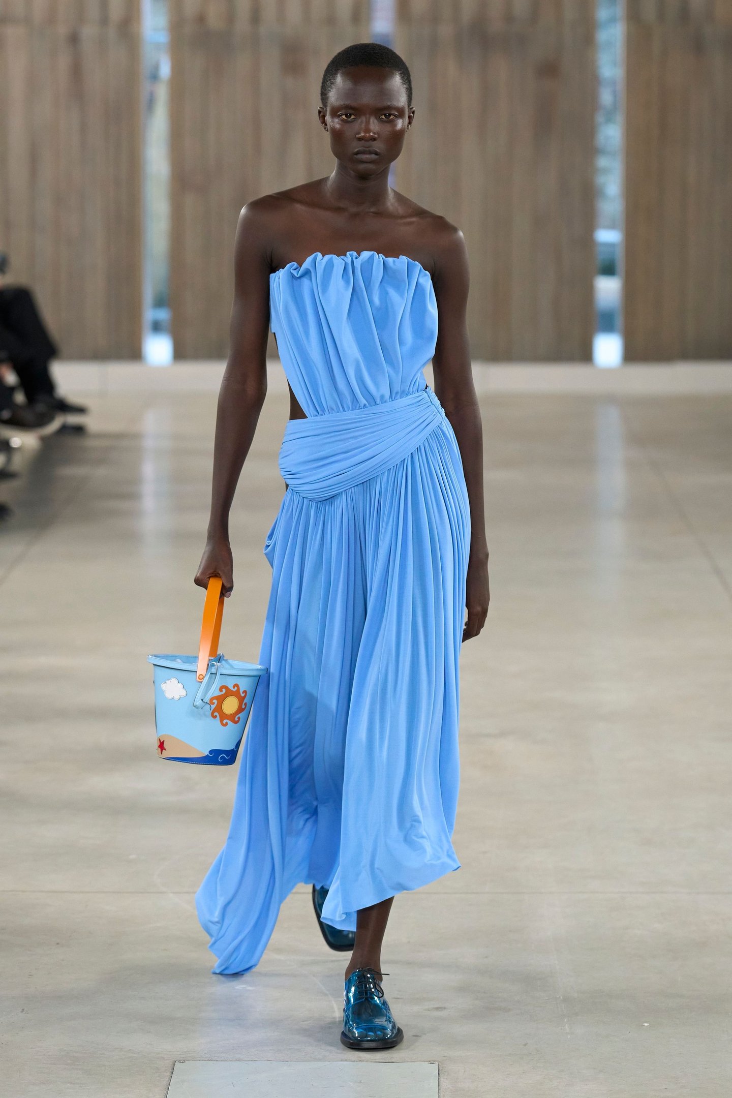 Modelo desfila vestido azul sem alças carregando balde decorado. Semana de Moda de Milão SS26.