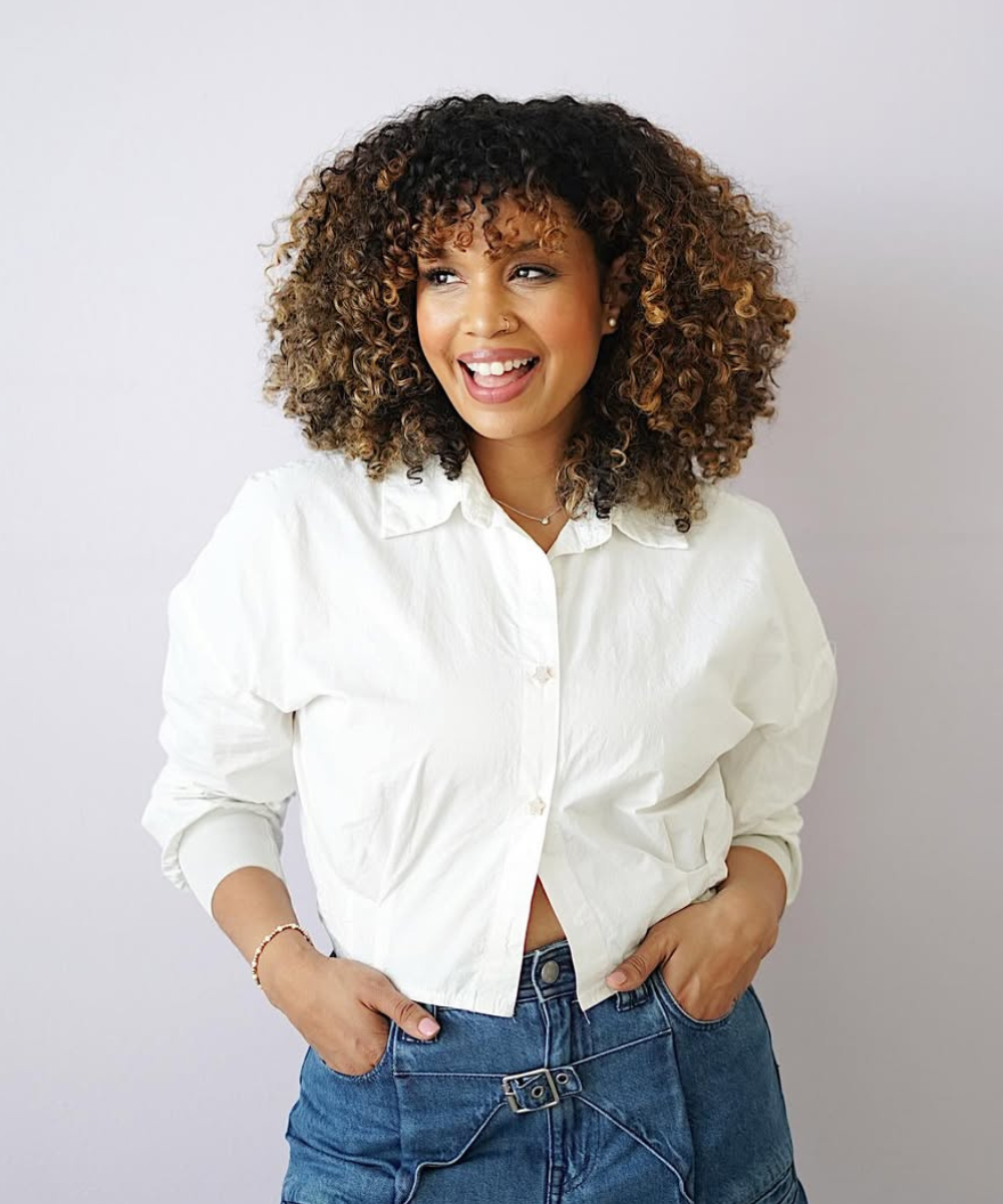 Pessoa sorrindo com cabelo cacheado, vestindo camisa branca e jeans. Cabelo vibrante sugere uso do melhor shampoo para queda.