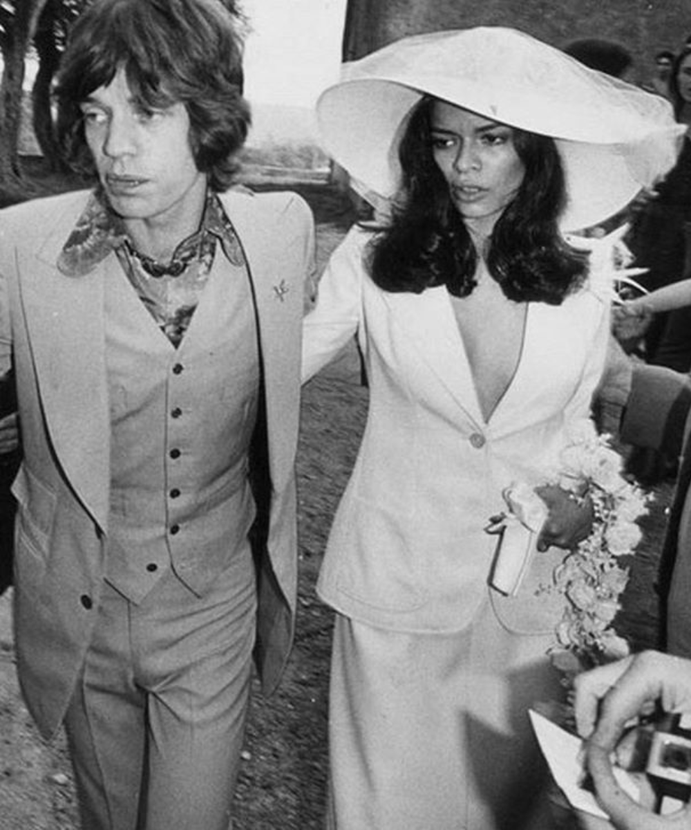 Homem e mulher caminhando, ela com traje branco e chapéu grande, um vestido não tradicional para a ocasião.