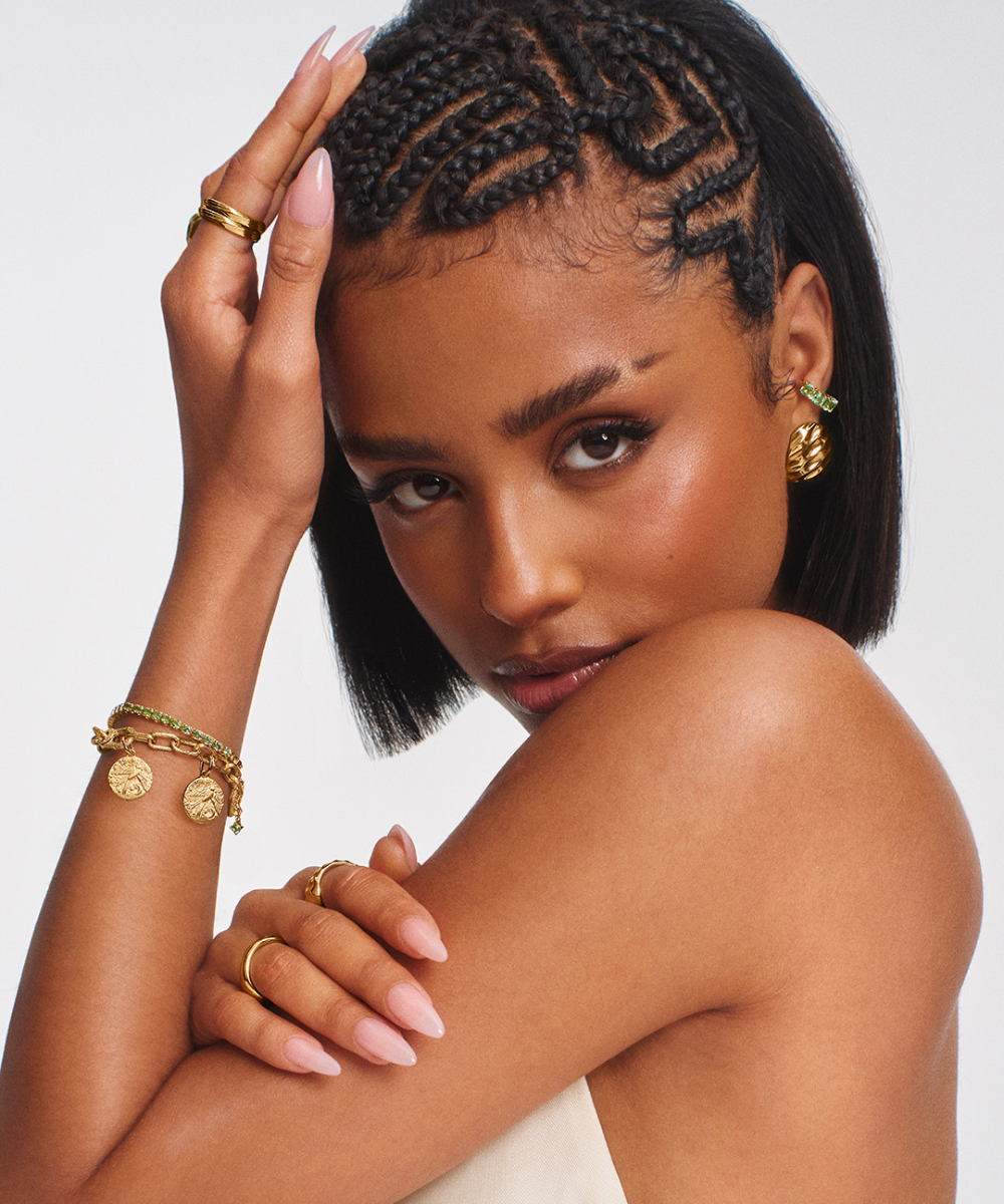 Mulher com tranças e pele morena usa joias douradas no braço e orelha. Ela exibe bracelete Pandora Talisman.