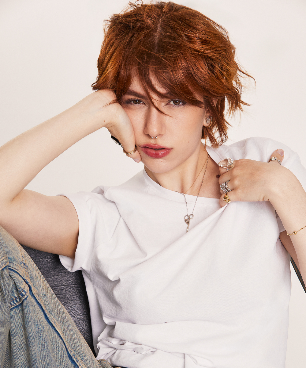 Uma pessoa com cabelo ruivo e expressão pensativa, usando camiseta branca e acessórios; Iguatemi Talks Fashion.