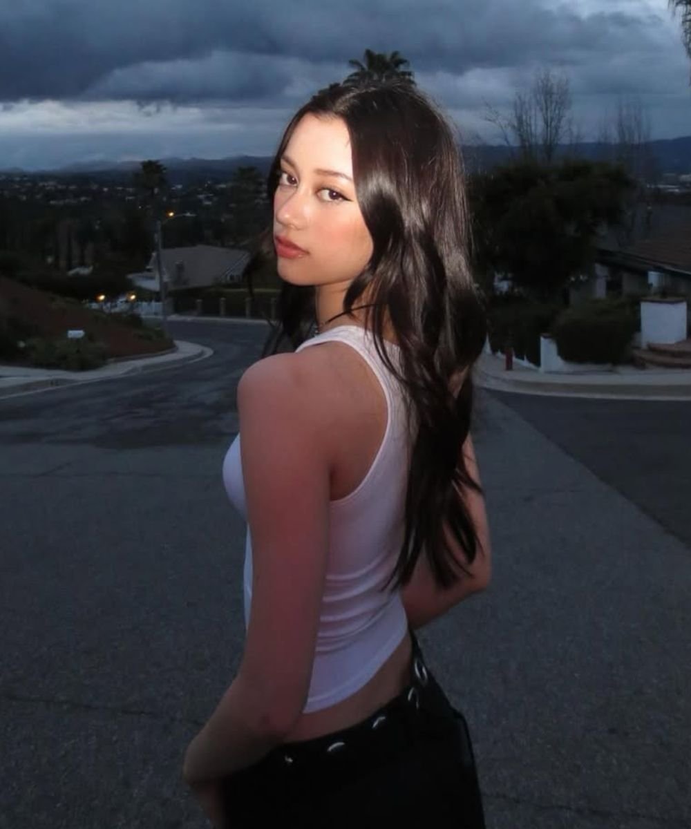 Mulher com cabelo ondulado usa regata branca, posando em rua sob céu nublado, possível uso de creme para cabelo ondulado