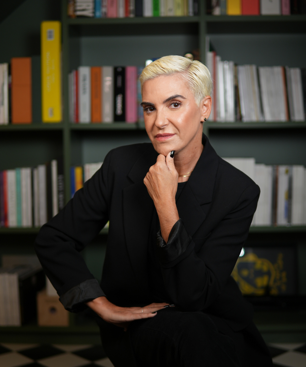 A imagem mostra uma pessoa de cabelos curtos e loiros, vestindo um terno preto, sentada em frente a uma estante de livros. Essa cena remete a um ambiente sofisticado, estilo Iguatemi Talks Fashion.