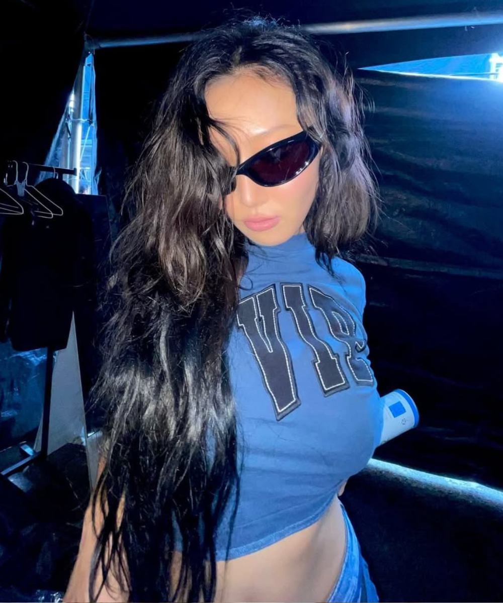 Uma pessoa de óculos de sol e cabelos longos veste camiseta azul estampada em um ambiente escuro. Fios saudáveis depois de usar o melhor shampoo para queda de cabelo