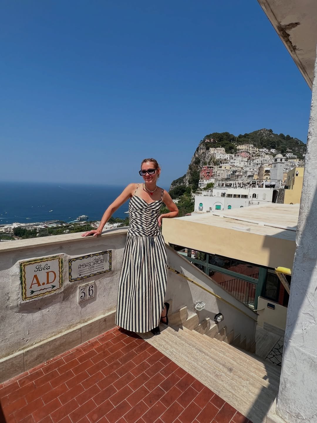 Mulher com vestido listrado preto e branco em varanda com vista para cidade costeira e mar sob céu azul claro.