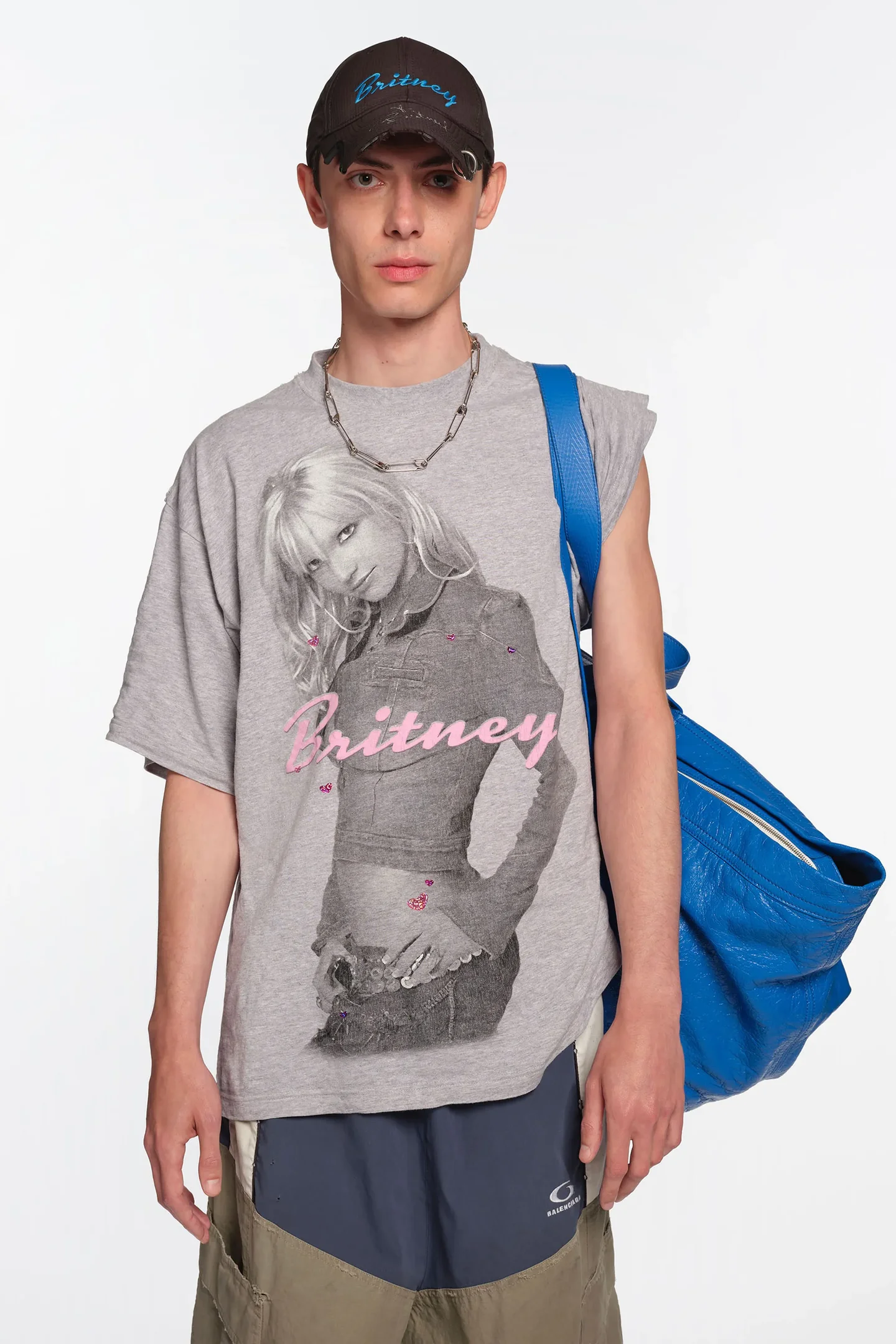 Jovem com graphic tee cinza estampada, carregando bolsa azul. Ele usa boné preto e colar prata.