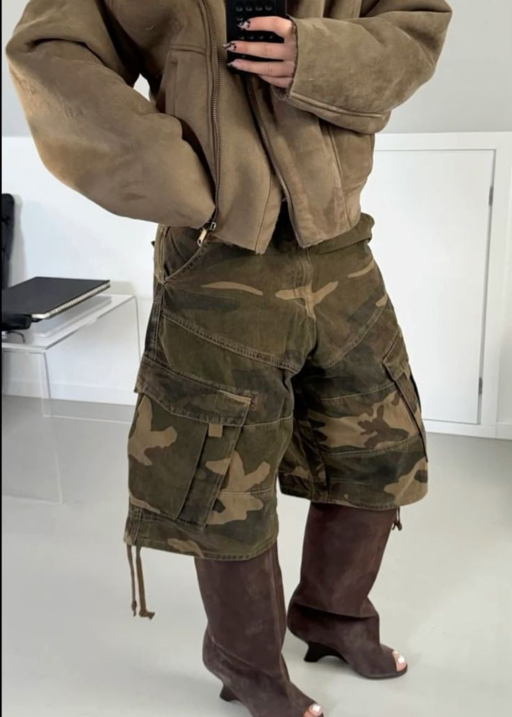 Pessoa usando jaqueta marrom, bermuda camuflada e botas marrons em um ambiente interno.