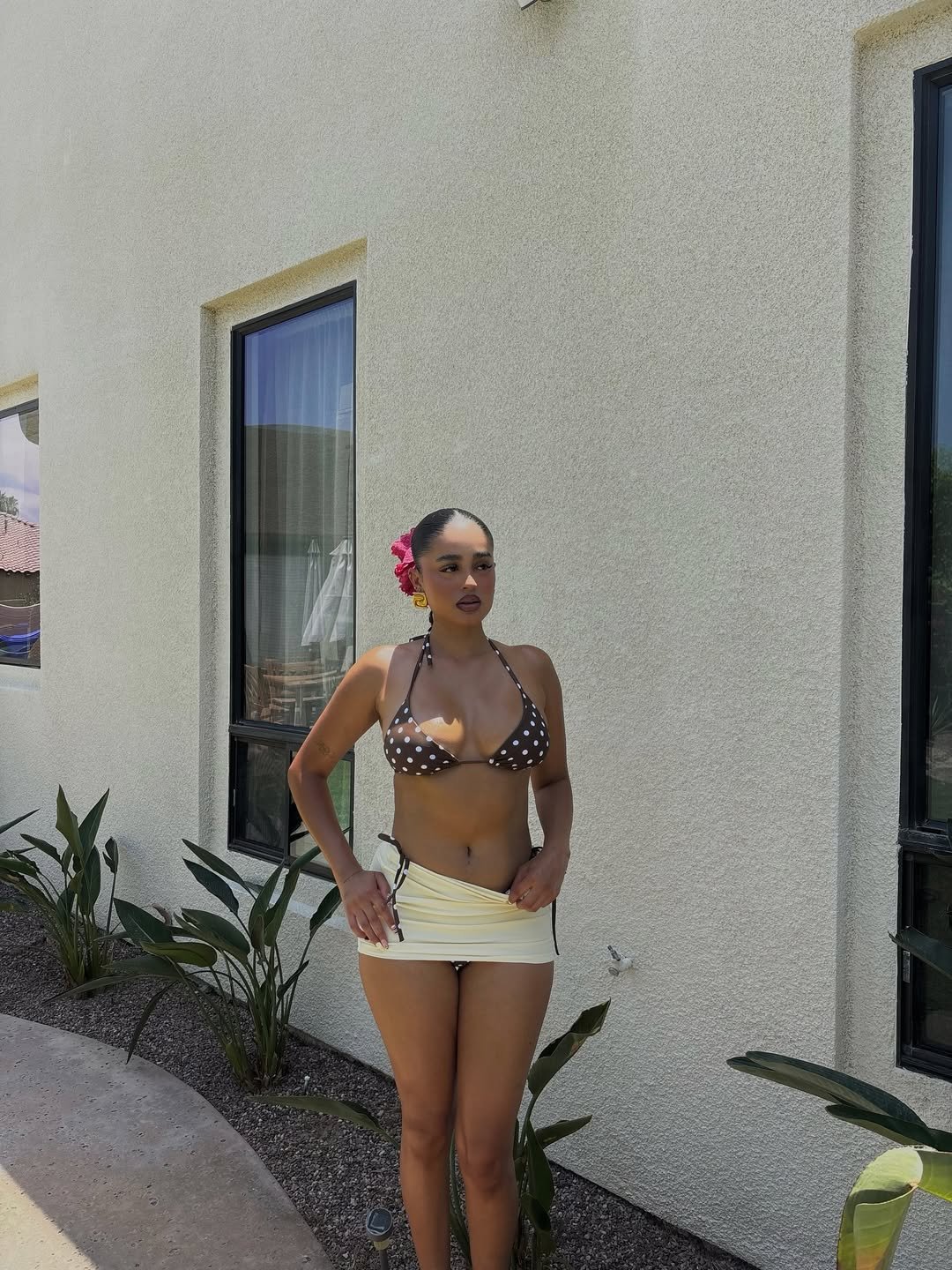 Pessoa posando ao ar livre, usando um biquíni da moda marrom de bolinhas com saia branca em local ensolarado.