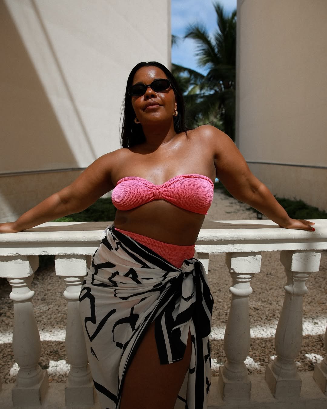 Pessoa usando biquíni da moda e óculos escuros, apoiada em uma varanda com palmeiras ao fundo.