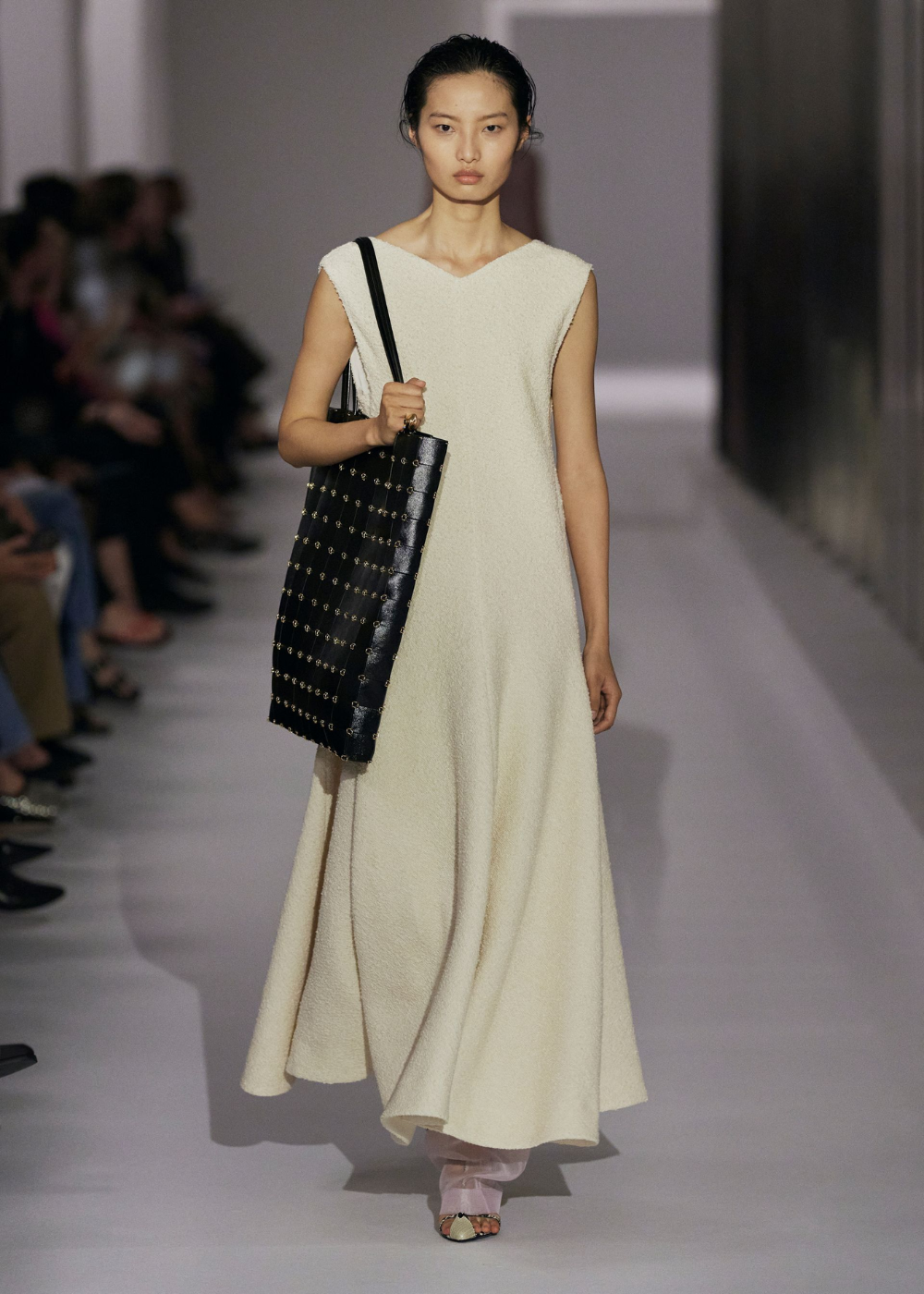 Desfile de moda com modelo usando vestido longo bege e bolsa de stud preta.