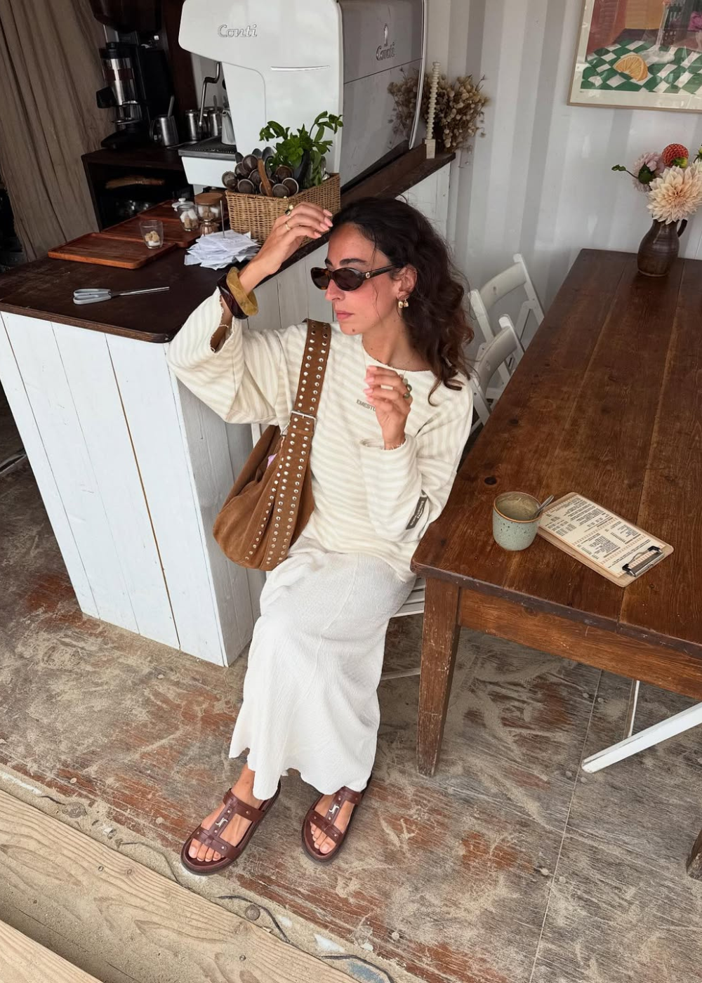 Mulher de óculos escuros segura uma bolsa marrom ao lado de uma mesa de madeira.