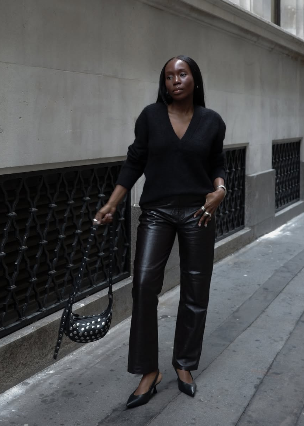 Mulher com suéter preto, calça de couro e bolsa de mão com detalhes brancos, caminhando em rua urbana.