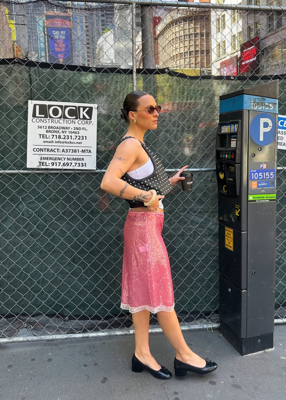 Pessoa de óculos escuros e bolsa de studs, segurando copo ao lado de um parquímetro.