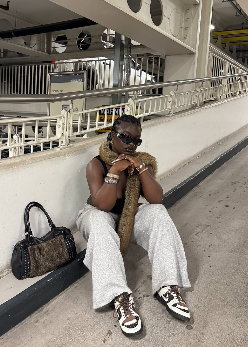 Pessoa sentada em garagem vestindo colete de pele e calça cinza, com bolsa de mão preta com detalhes.