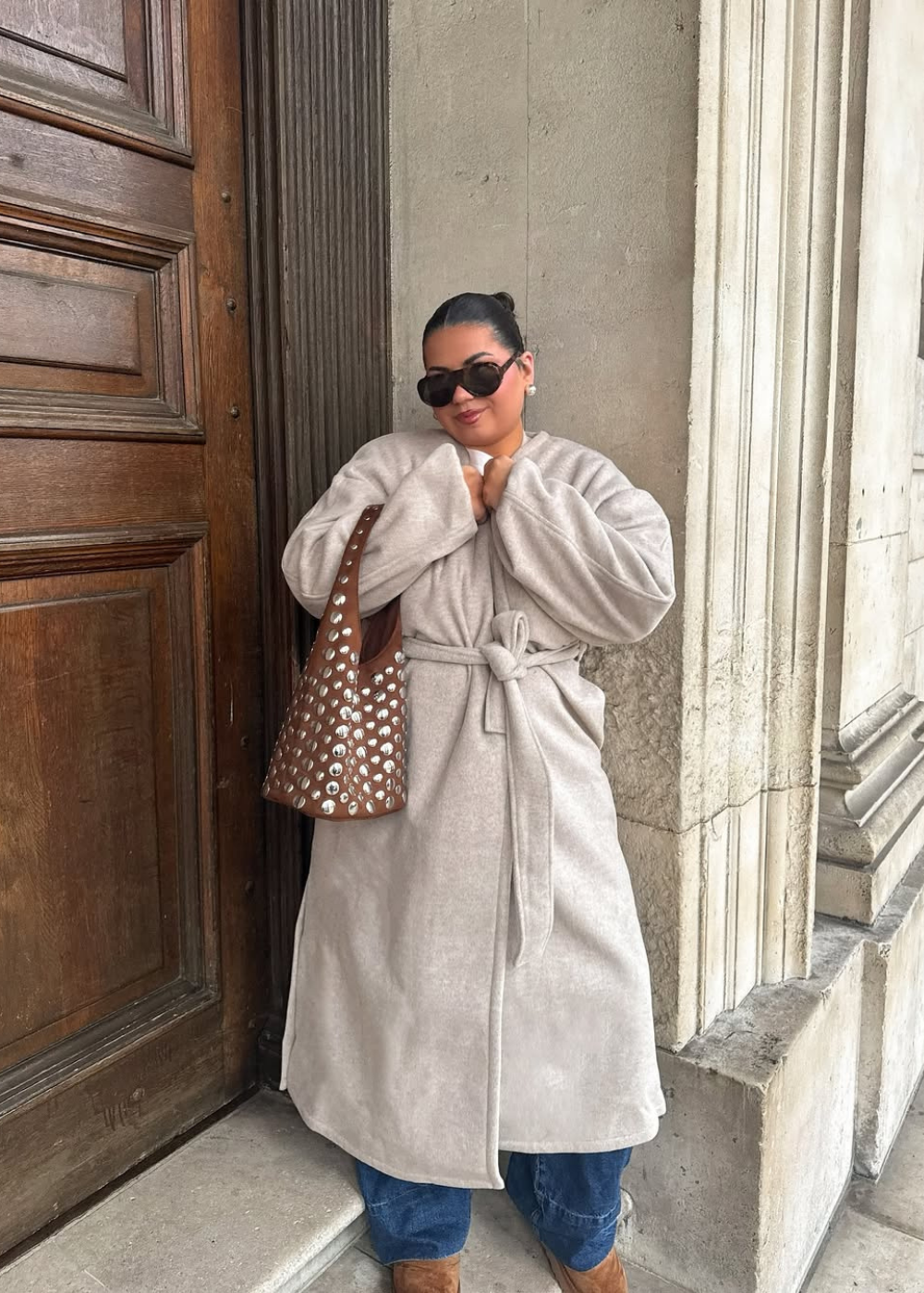 Pessoa com casaco bege, bolsa de tachas brilhantes, óculos escuros, encostada em parede clássica.