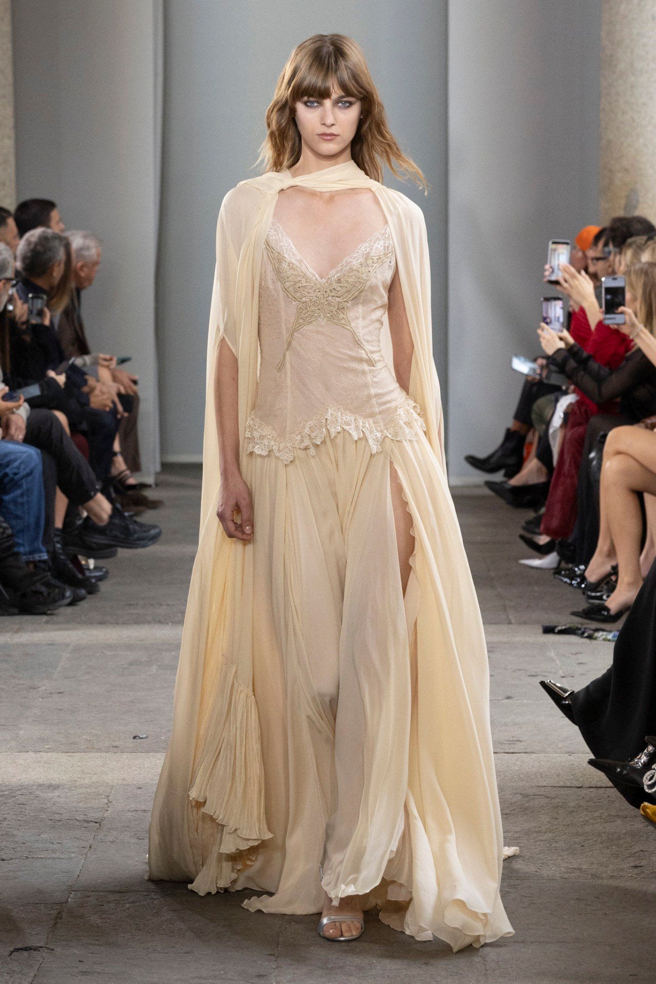 Modelo desfila com vestido elegante em tom creme durante a Milan Fashion Week SS26, cercada por uma audiência atenta.