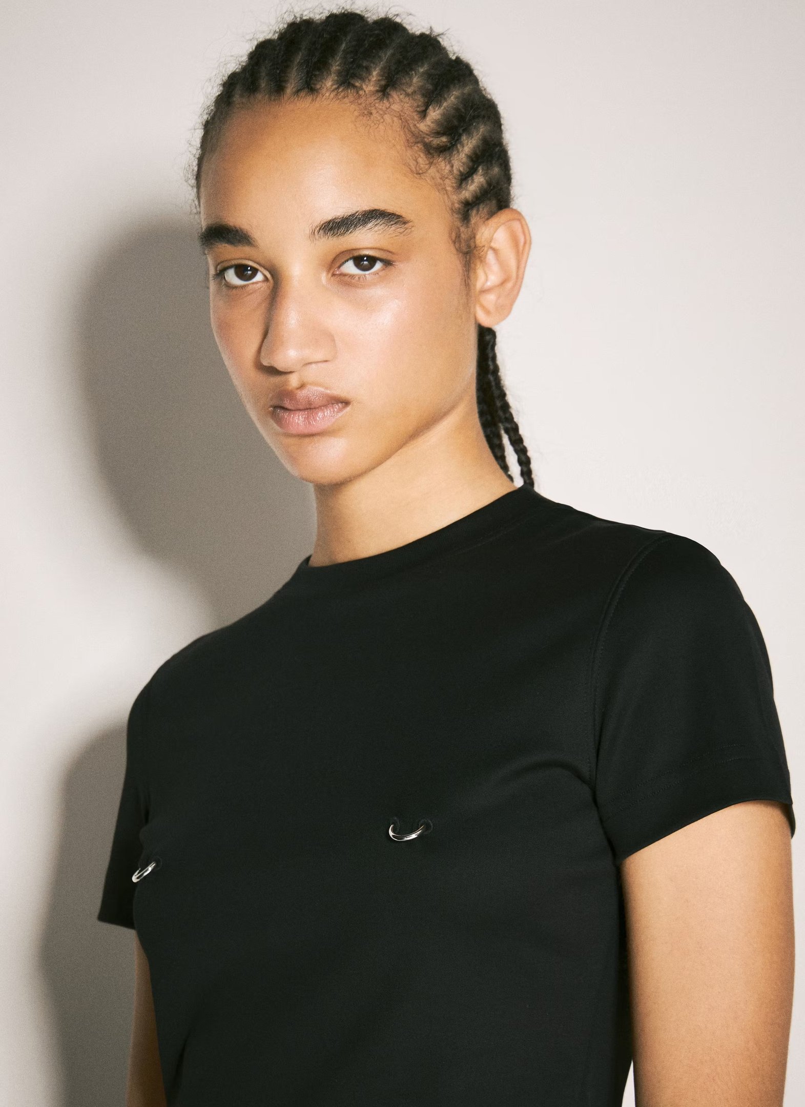 Jovem com cabelo trançado, usando camiseta preta com anéis metálicos, refletindo a moda dos piercings.