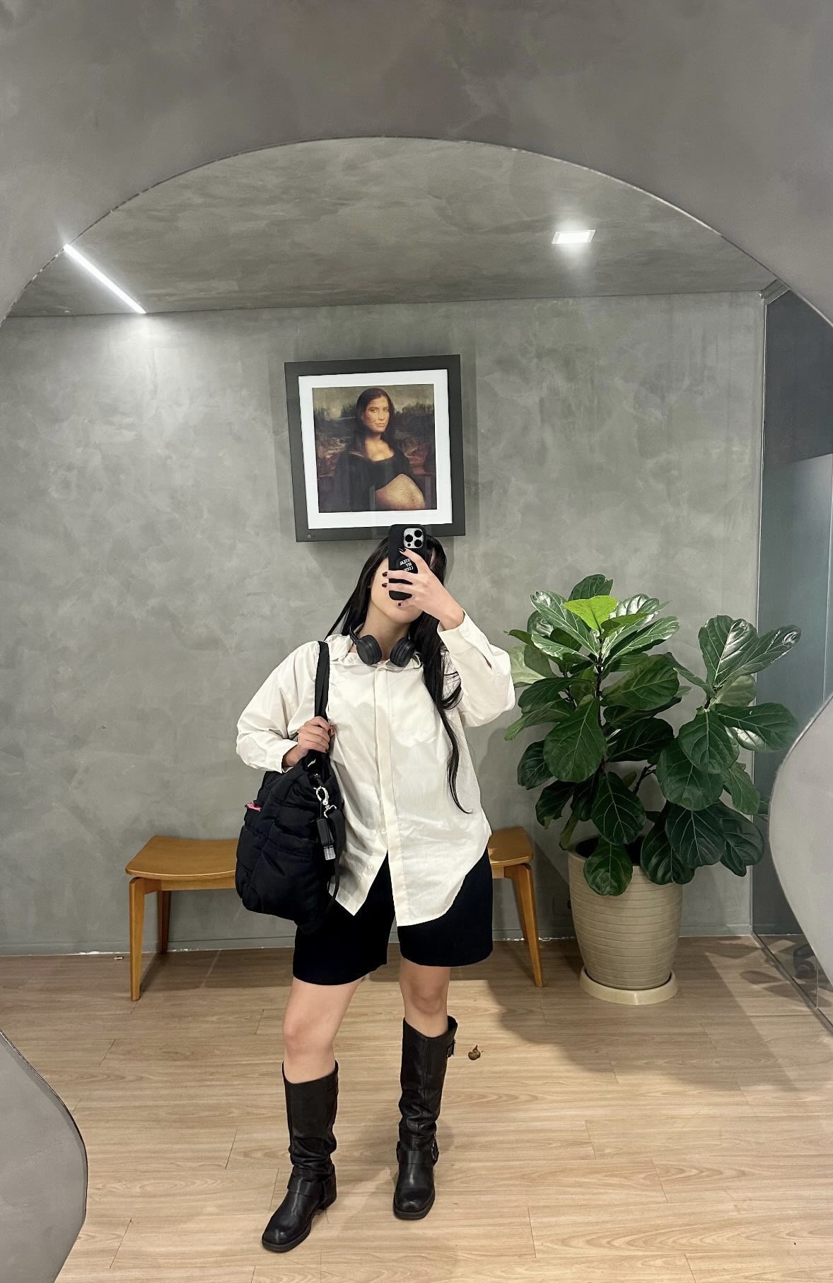 Pessoa vestida com camisa branca e botas, segurando uma bolsa. Há um quadro e uma planta ao fundo.
