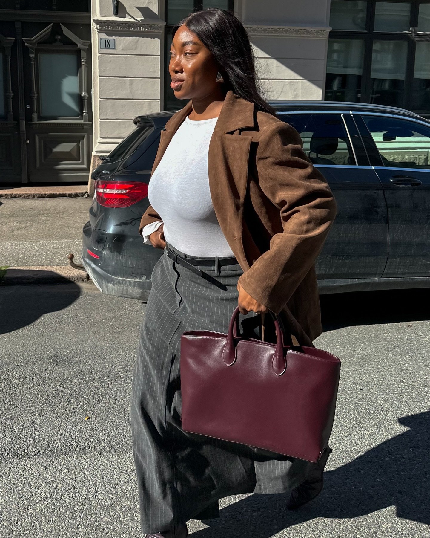Mulher caminhando com bolsa marrom de couro em uma rua ensolarada, usando casaco marrom e calças risca de giz.