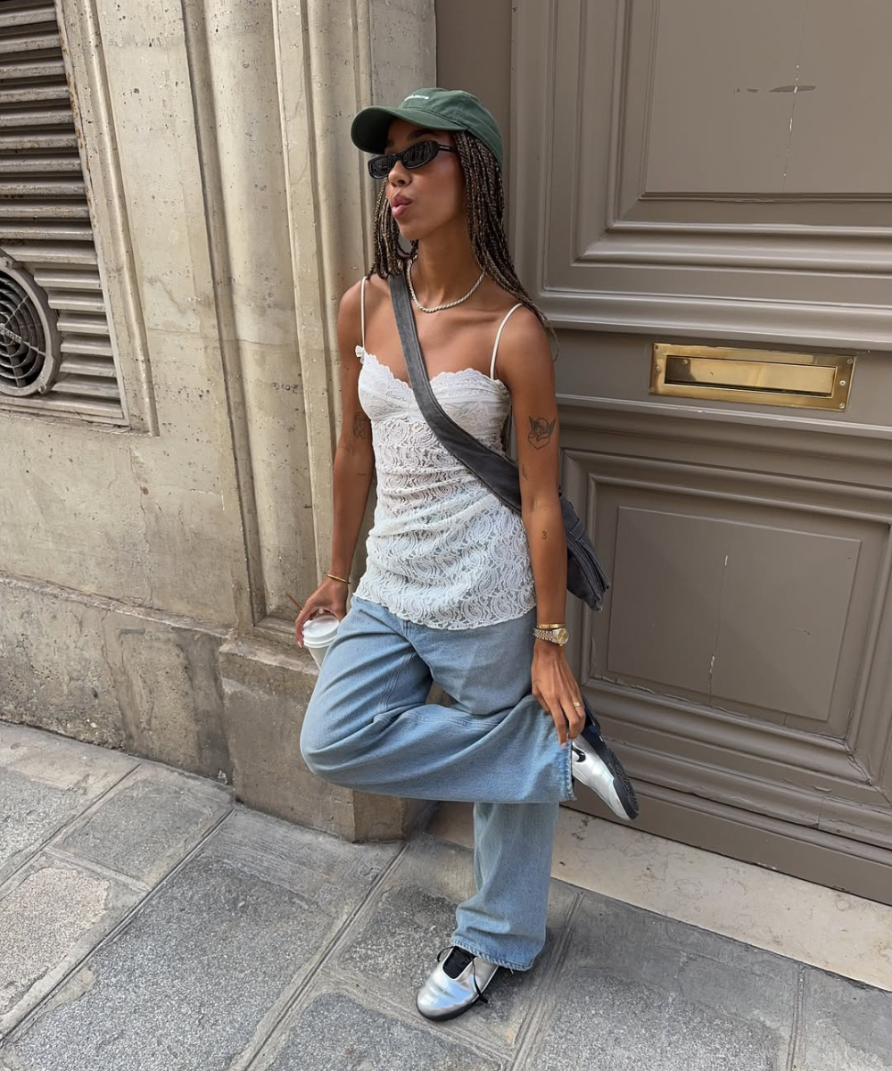 Pessoa com boné verde e óculos de sol, usando blusa de renda branca e calça jeans larga, encostada em uma parede.