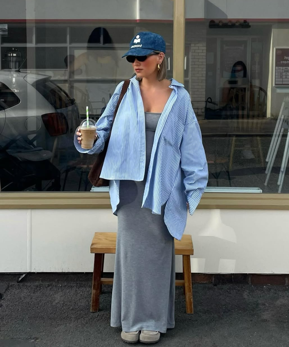 Mulher com boné azul e óculos de sol, usando camisa listrada sobre vestido cinza, segura bebida em frente a uma janela.