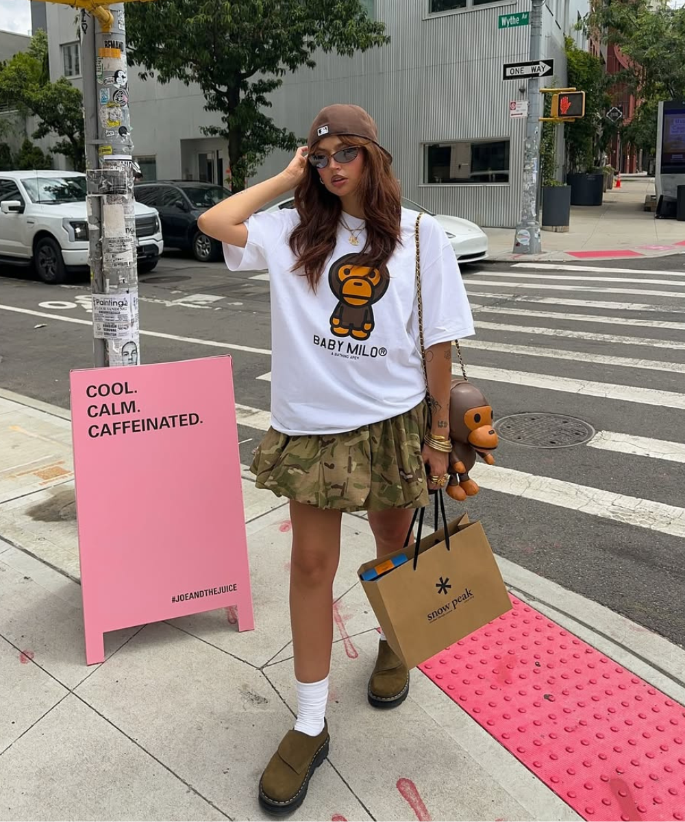 Pessoa com boné marrom e óculos de sol, usando camiseta estampada e saia camuflada em calçada urbana.